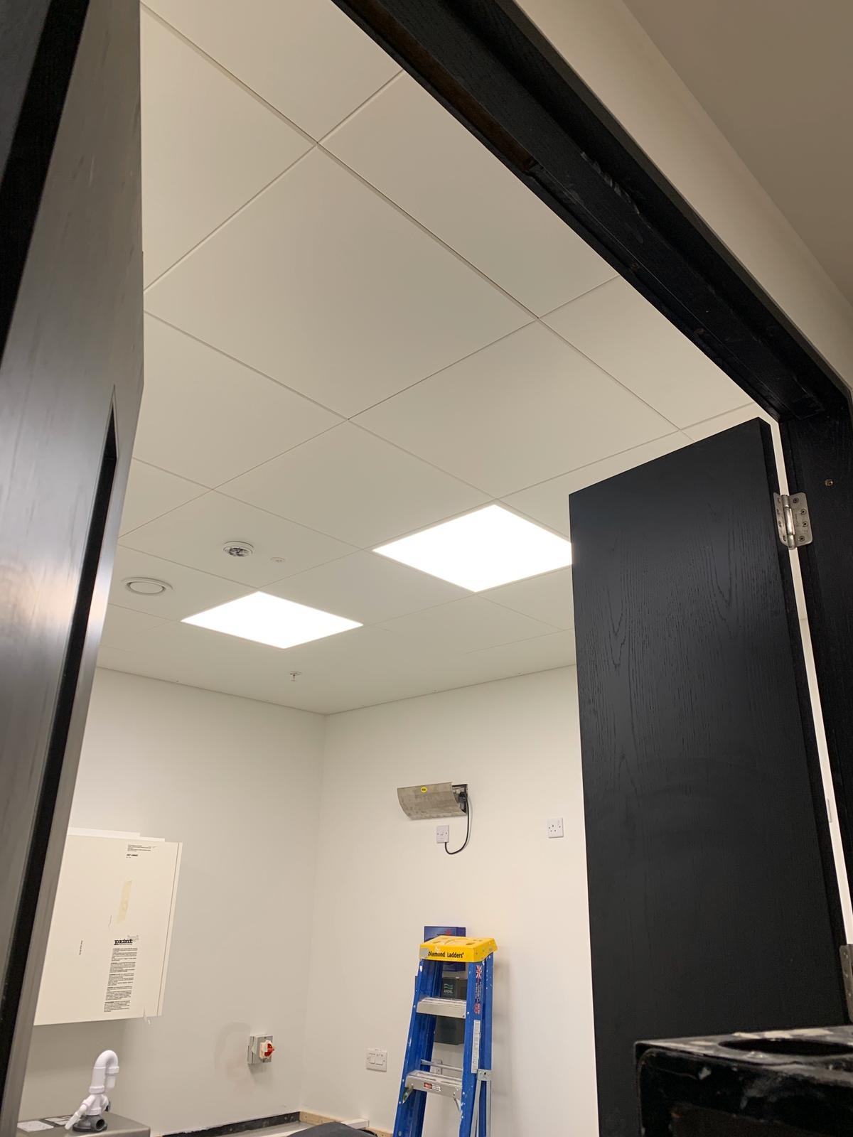 Widok przez czarną futrynę drzwi na wnętrze z białym sufitem podwieszanym z kwadratowymi panelami i wbudowanym oświetleniem, niebieska drabina stoi przy ścianie z widocznymi gniazdkami...