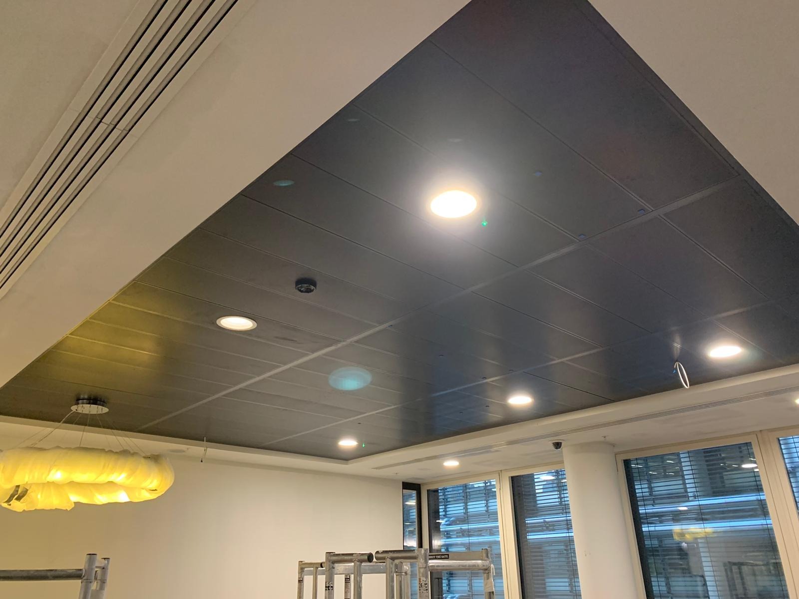 Metalowy sufit podwieszany w biurze z wbudowanym oświetleniem punktowym, widoczne kratki wentylacyjne i schodki malarskie.