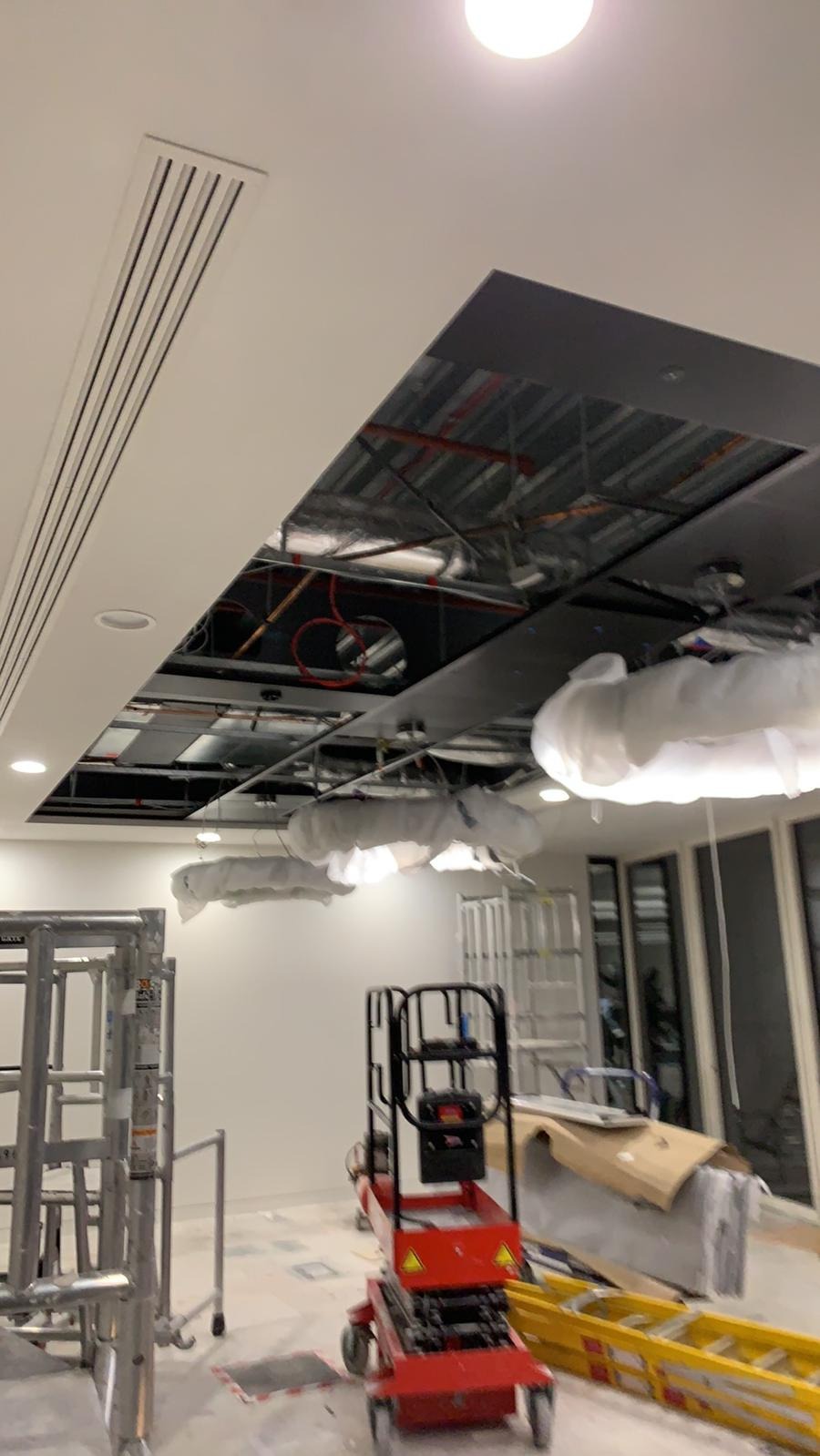 Montaż sufitu podwieszanego z widoczną instalacją, oświetleniem i kratką wentylacyjną, z użyciem podnośnika nożycowego i drabiny w trakcie remontu biura.