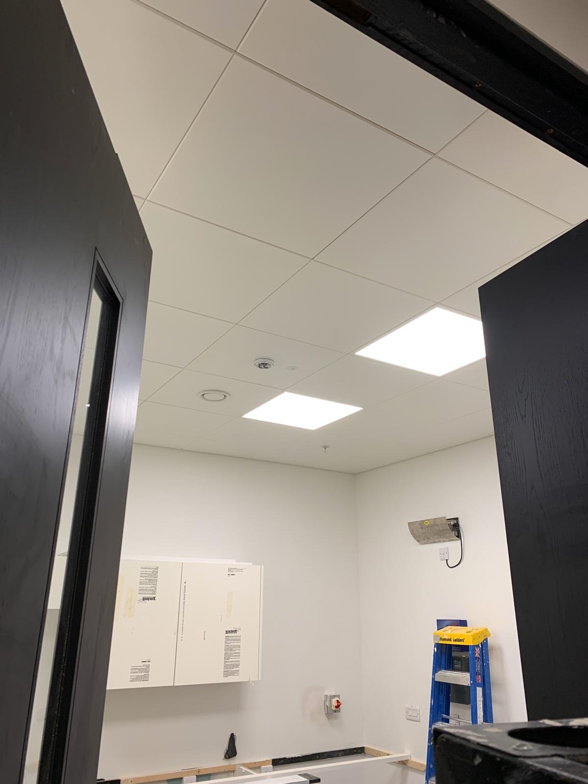 Widok z dołu na sufit modułowy z kwadratowymi panelami i wbudowanym oświetleniem LED w pomieszczeniu w trakcie remontu, widoczne drzwi po bokach, szafki na ścianie i drabina