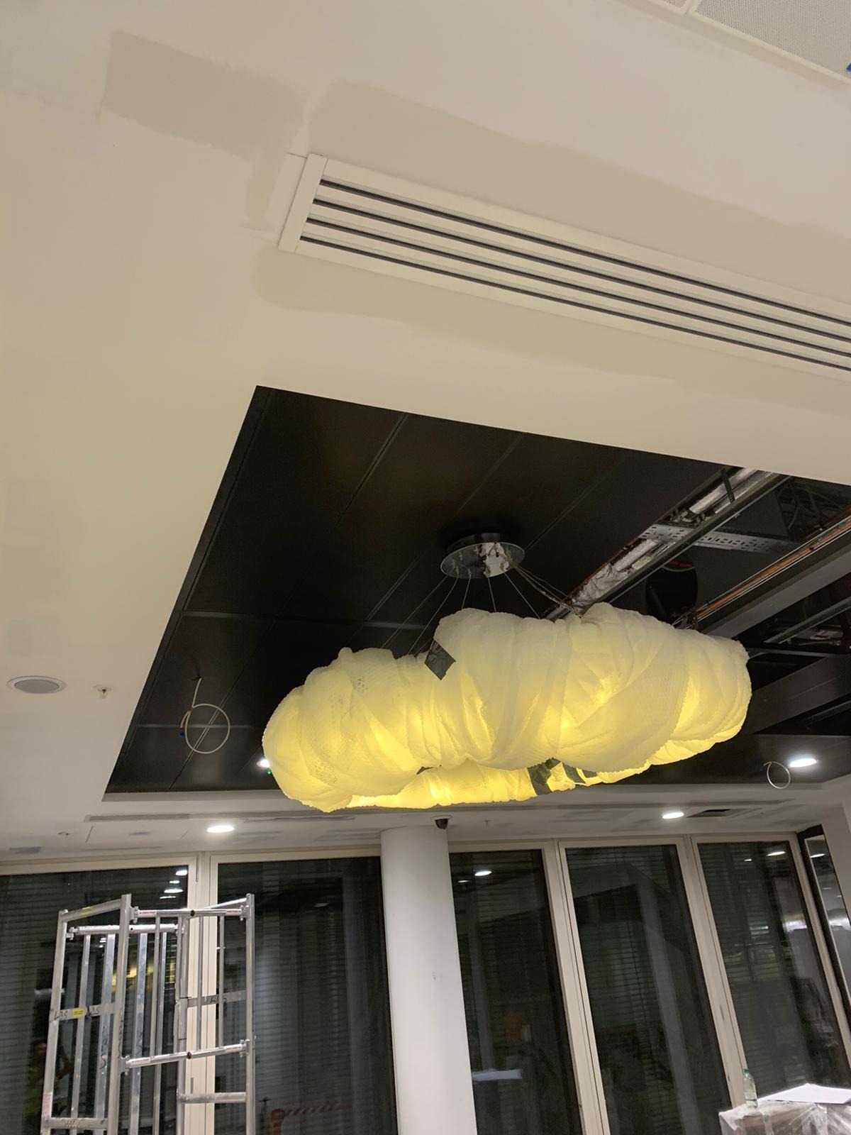 Niedokończone prace sufitowe z widocznymi elementami instalacji elektrycznej i dekoracyjną lampą w kształcie chmury, drabina w tle, fragment wentylacji sufitowej z widocznymi śladami szpachlowania.