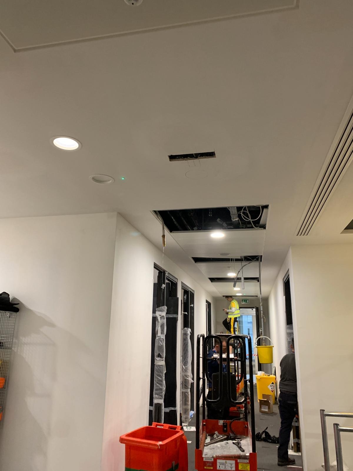 Korytarz biurowy w trakcie prac remontowych sufitu podwieszanego, widoczny pracownik na podnośniku koszowym, otwarte przestrzenie w suficie ukazujące instalacje, zarysowane miejsca pod nowe...