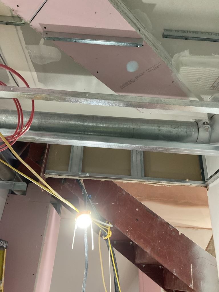 Montaż metalowego stelażu pod sufitem z płyt gipsowo-kartonowych, widoczne przewody elektryczne i rura wentylacyjna, oświetlenie robocze.