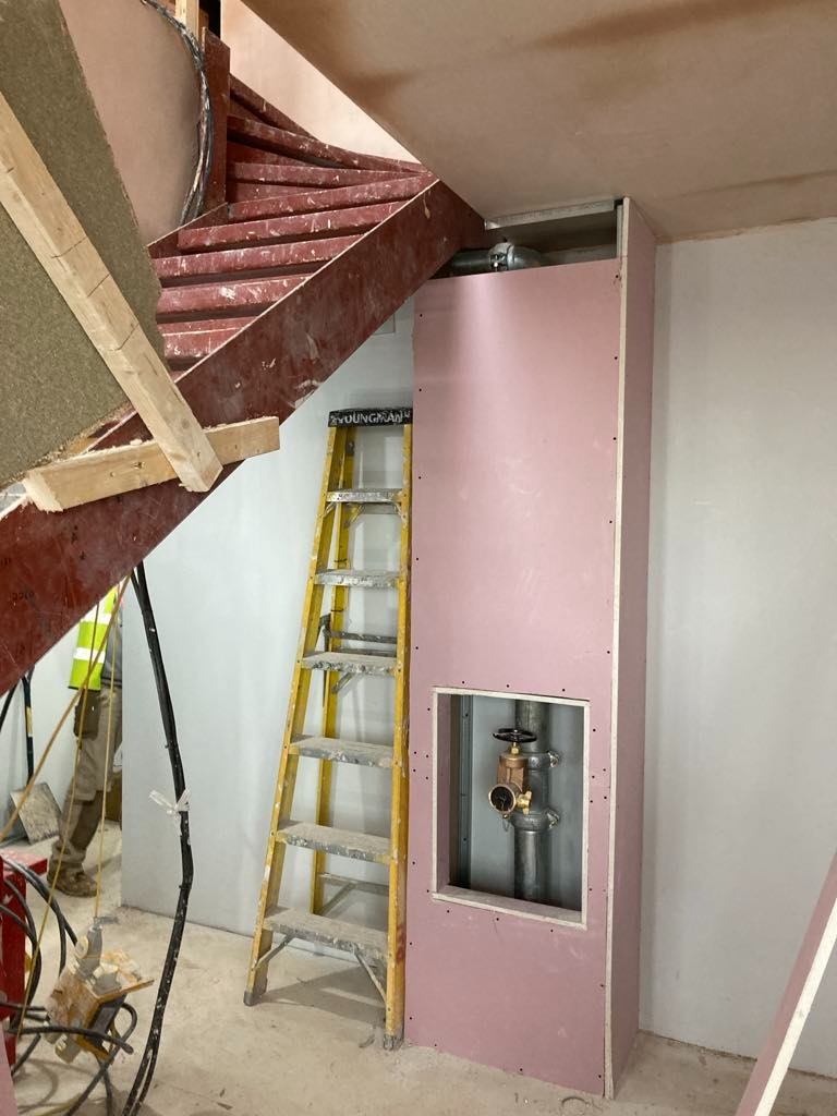 Montaż obudowy z różowej płyty gipsowej wokół rur z zaworem, widoczna żółta drabina i fragment schodów spiralnych w surowym stanie.