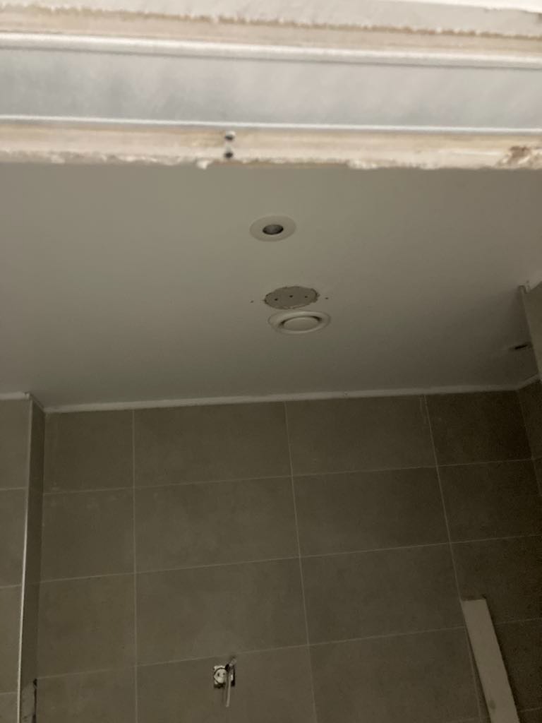 Niedokończony sufit podwieszany z widocznymi otworami na oświetlenie punktowe i szkieletem konstrukcji, szare płytki na ścianie, widoczny gniazdko elektryczne.