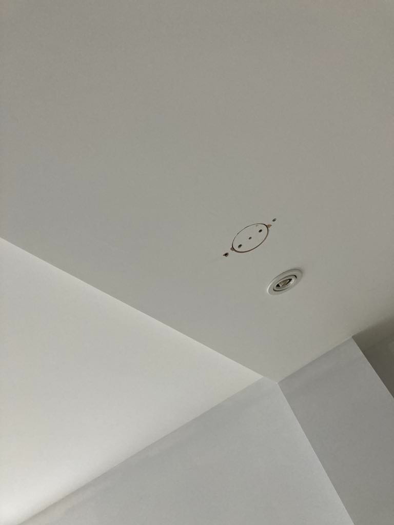 Biały sufit z widocznym okrągłym otworem z trzema małymi otworami w środku, obok oprawa oświetleniowa typu downlight.