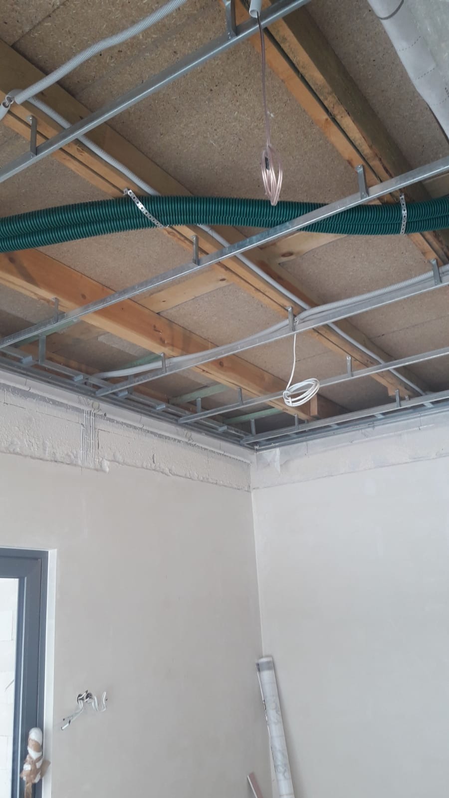 Sufit podwieszany w trakcie montażu: widoczne profile metalowe, drewniane elementy konstrukcyjne, zielona rura karbowana przymocowana do sufitu, częściowo wykończone ściany, wystające przewody...