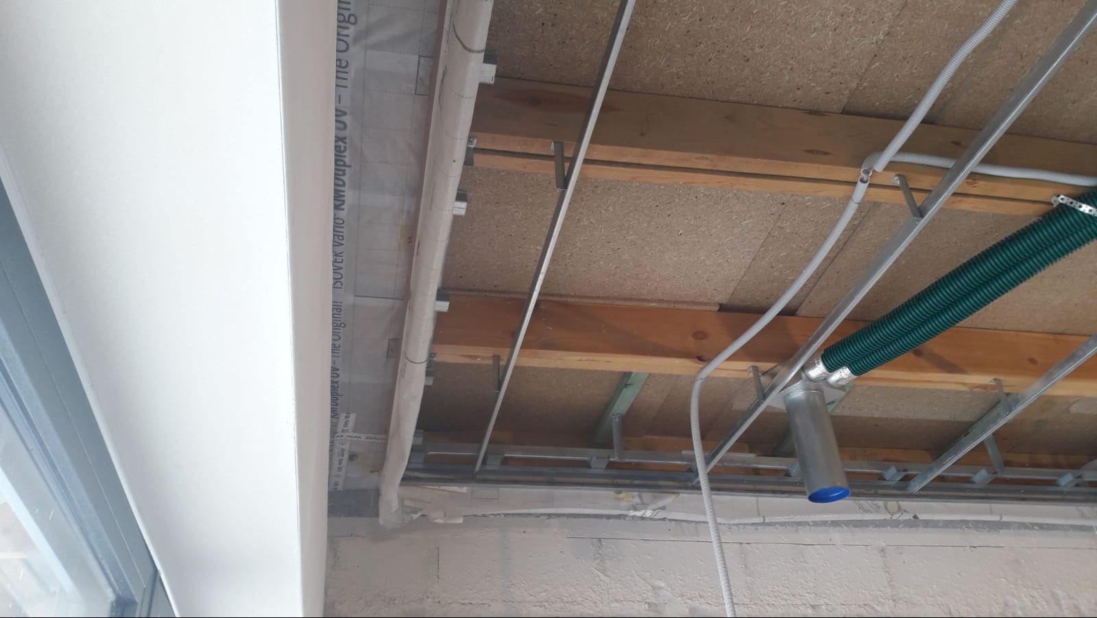 Sufit poddasza w trakcie prac: widoczne drewniane belki, metalowe profile, izolacja z folii paroizolacyjnej ISOVER, przewody elektryczne i zielony przewód wentylacyjny z niebieską zaślepką.