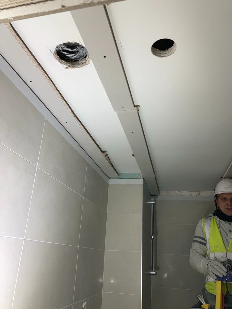 Montaż płyty karton gips na suficie w łazience z wyciętymi otworami na oświetlenie, widoczny glazurnik w kasku i kamizelce odblaskowej.