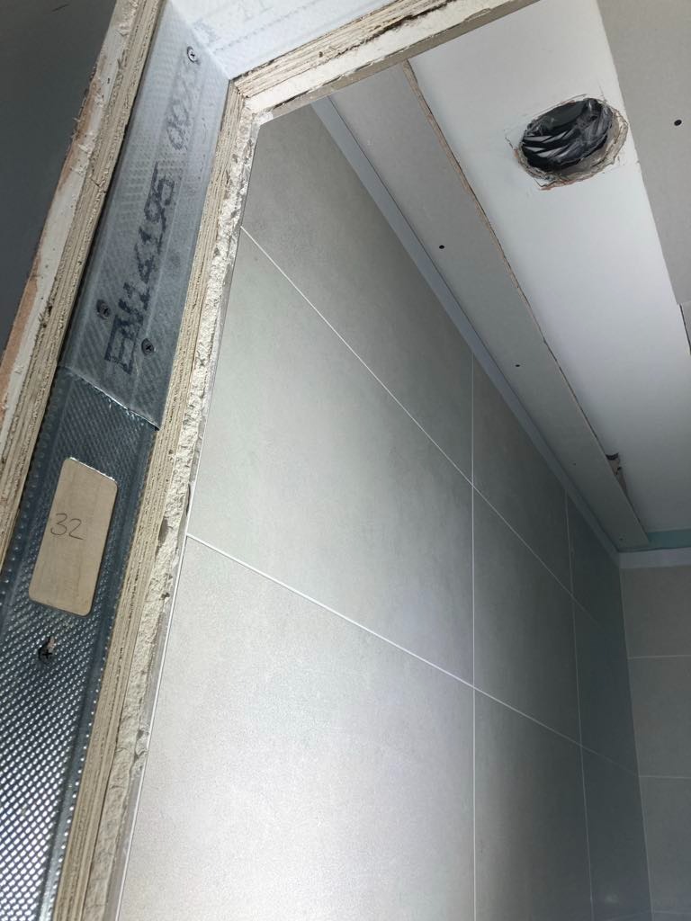 Wnętrze w trakcie wykańczania: widok na szkielet metalowy przy otworze drzwiowym, fragment ściany wyłożonej szarymi płytkami oraz otwór w suficie z zabezpieczonymi kablami.