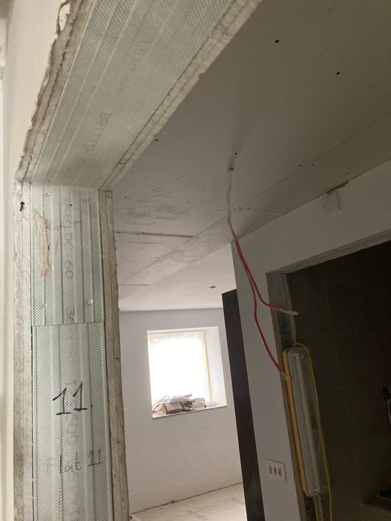 Fragment ściany z zamontowanymi profilami i częściowo położoną płytą, widoczne przewody elektryczne, w tle okno z widokiem na plac budowy.