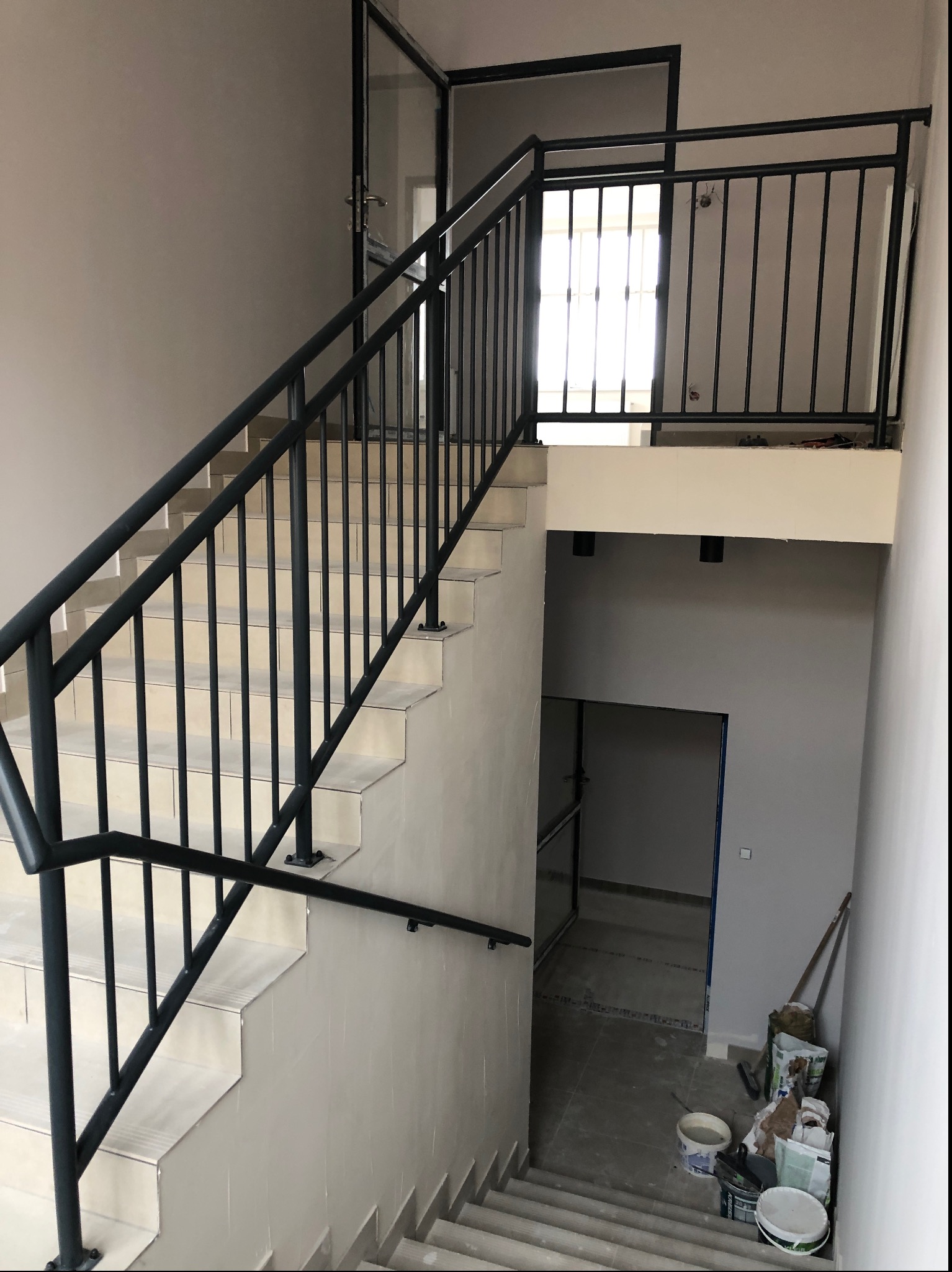 Klatka schodowa w trakcie wykańczania z czarną metalową balustradą, widoczne materiały budowlane i otwarte drzwi na półpiętrze.