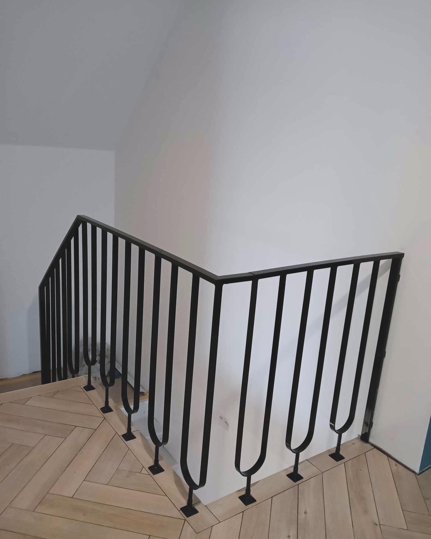 Nowoczesna, czarna balustrada schodowa z pionowymi prętami o zaokrąglonych zakończeniach, zamontowana na drewnianych schodach ułożonych w jodełkę, na tle białej ściany.