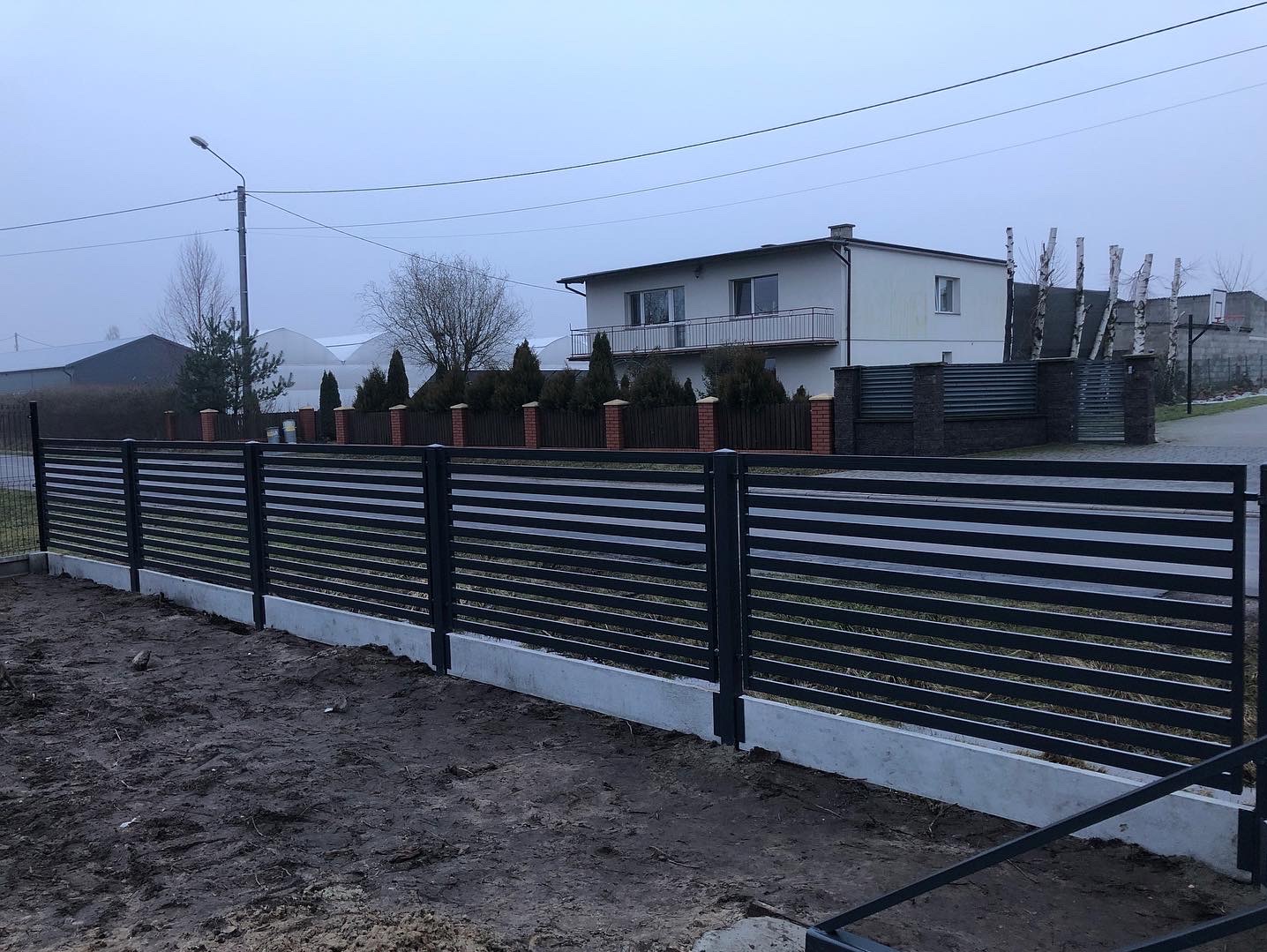 Nowoczesne ogrodzenie panelowe z poziomych, czarnych profili na betonowej podmurówce, widoczne na tle domu z balkonem i pochmurnego nieba.