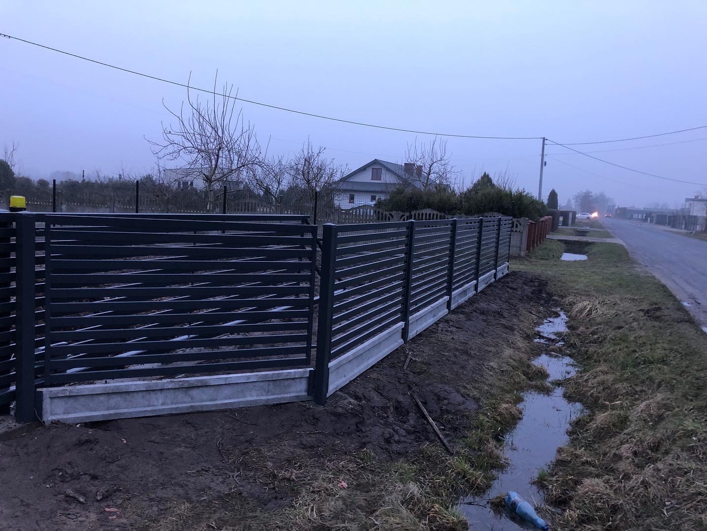 Nowoczesne ogrodzenie z poziomych, metalowych paneli w kolorze grafitowym, osadzone na betonowej podmurówce, widok z boku w pochmurny dzień, z widocznym rowem melioracyjnym i domem w tle.