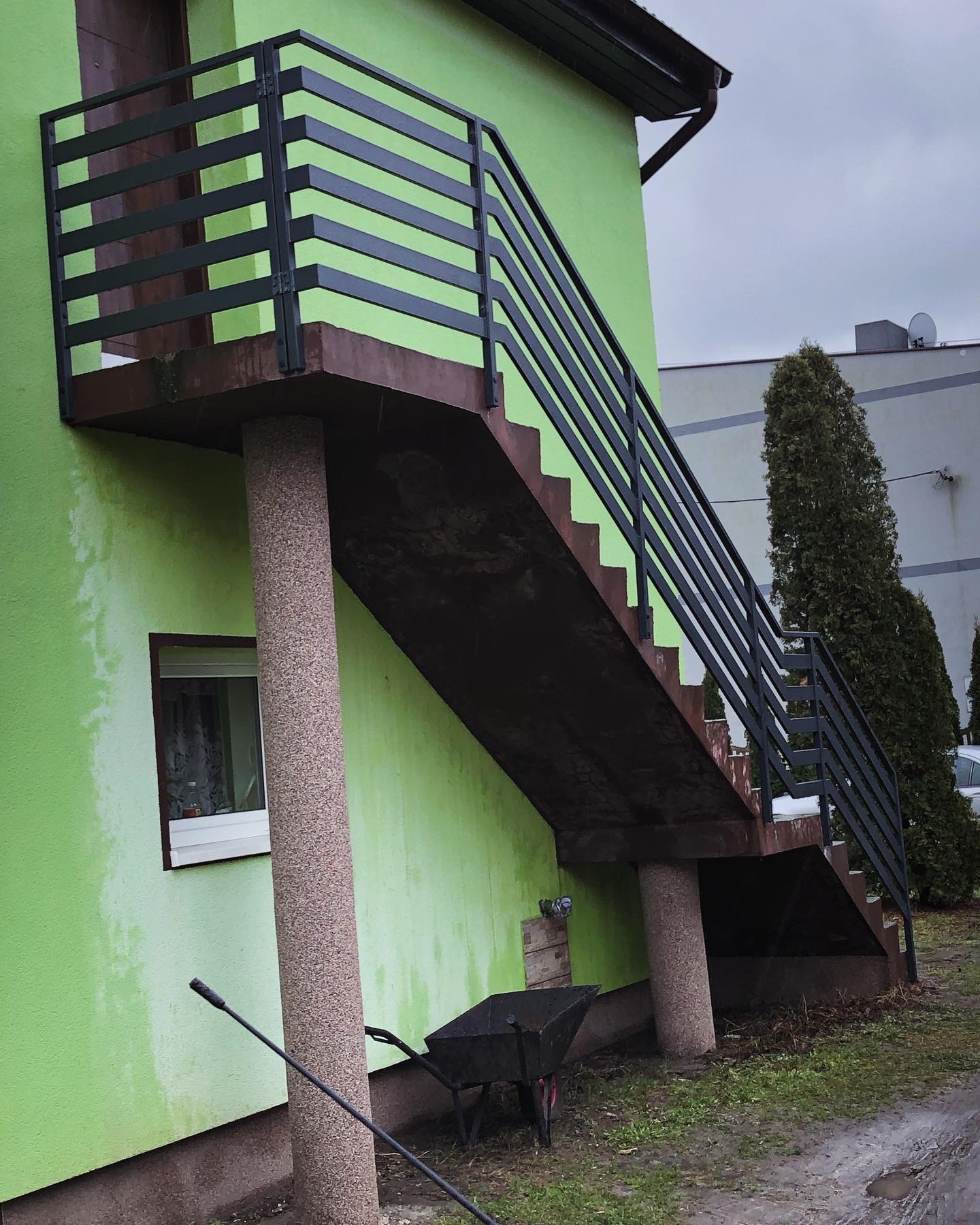 Zewnętrzne schody z czarną, metalową balustradą z poziomych prętów, prowadzące na balkon domu o zielonej elewacji, wsparte na dwóch kolumnach z fakturą kamienia, w tle wózek budowlany i zieleń.