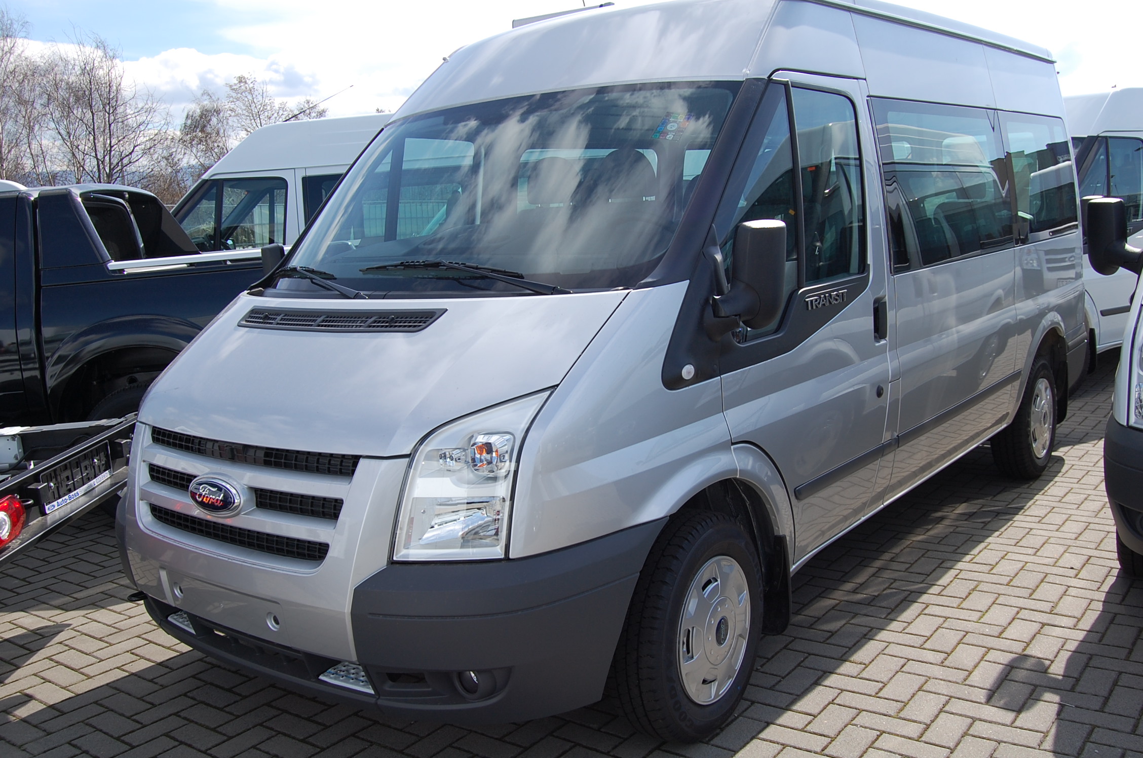 Srebrny Ford Transit, widok z przodu, zaparkowany na kostce brukowej, z odbiciem chmur w szybie.