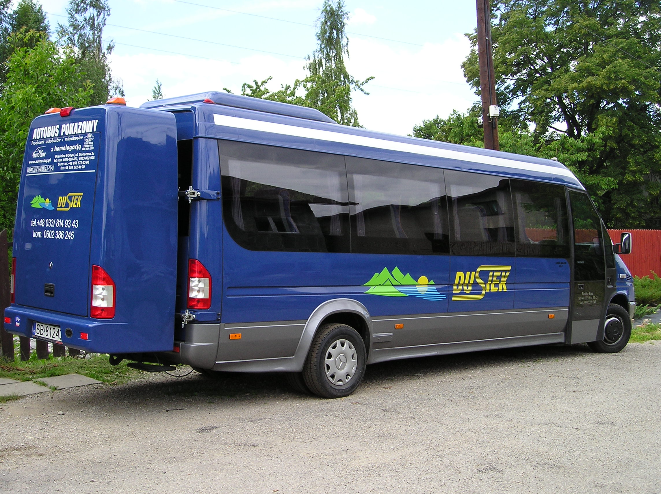 Niebieski bus z otwartymi drzwiami bagażnika, z logo firmy Duś-jek i napisem 'Autobus pokazowy' na tylnej klapie, zaparkowany na żwirowej nawierzchni.