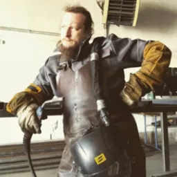 Mężczyzna w kombinezonie roboczym i skórzanym fartuchu, z maską spawalniczą na ramieniu i uchwytem spawalniczym w dłoni, w warsztacie.