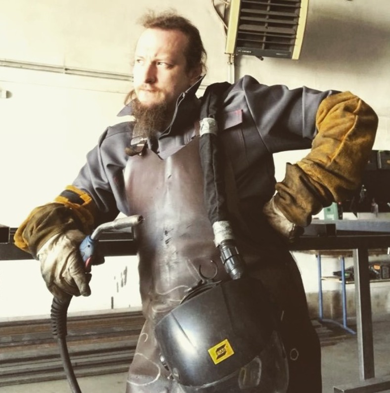 Mężczyzna w kombinezonie roboczym i skórzanym fartuchu, z maską spawalniczą na ramieniu i uchwytem spawalniczym w dłoni, w warsztacie.
