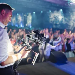 Kamerzysta obsługuje profesjonalny stabilizator obrazu Ronin z zamontowaną kamerą na scenie podczas koncertu, w tle rozświetlony tłum.