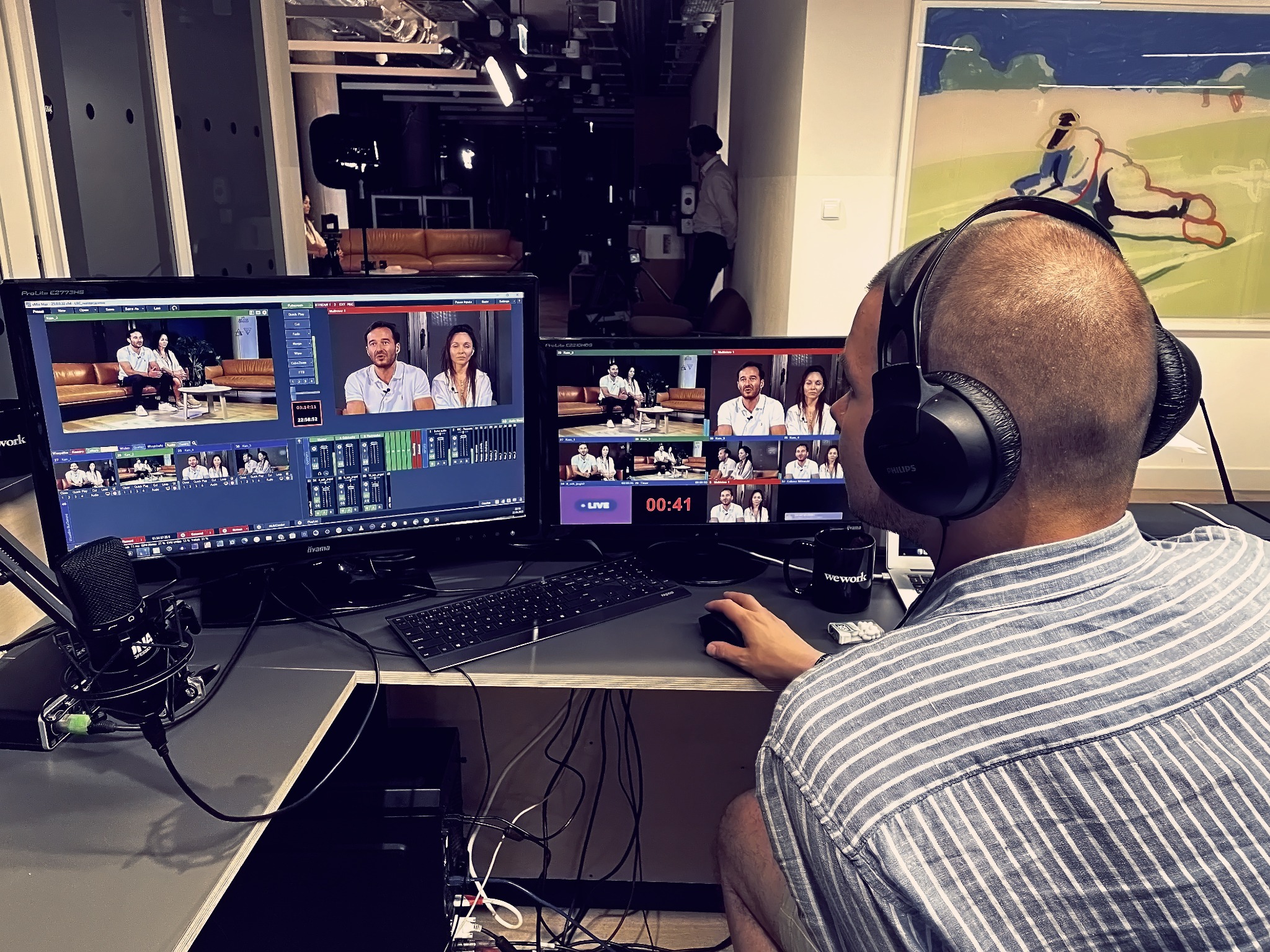 Operator w słuchawkach pracuje przy dwóch monitorach z podglądem nagrania rozmowy, mikrofon studyjny po lewej stronie, widoczny fragment biura z oświetleniem i obrazem na ścianie.