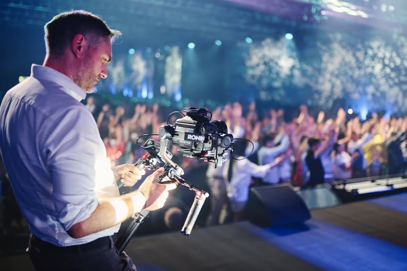 Kamerzysta obsługuje profesjonalny stabilizator obrazu Ronin z zamontowaną kamerą na scenie podczas koncertu, w tle rozświetlony tłum.