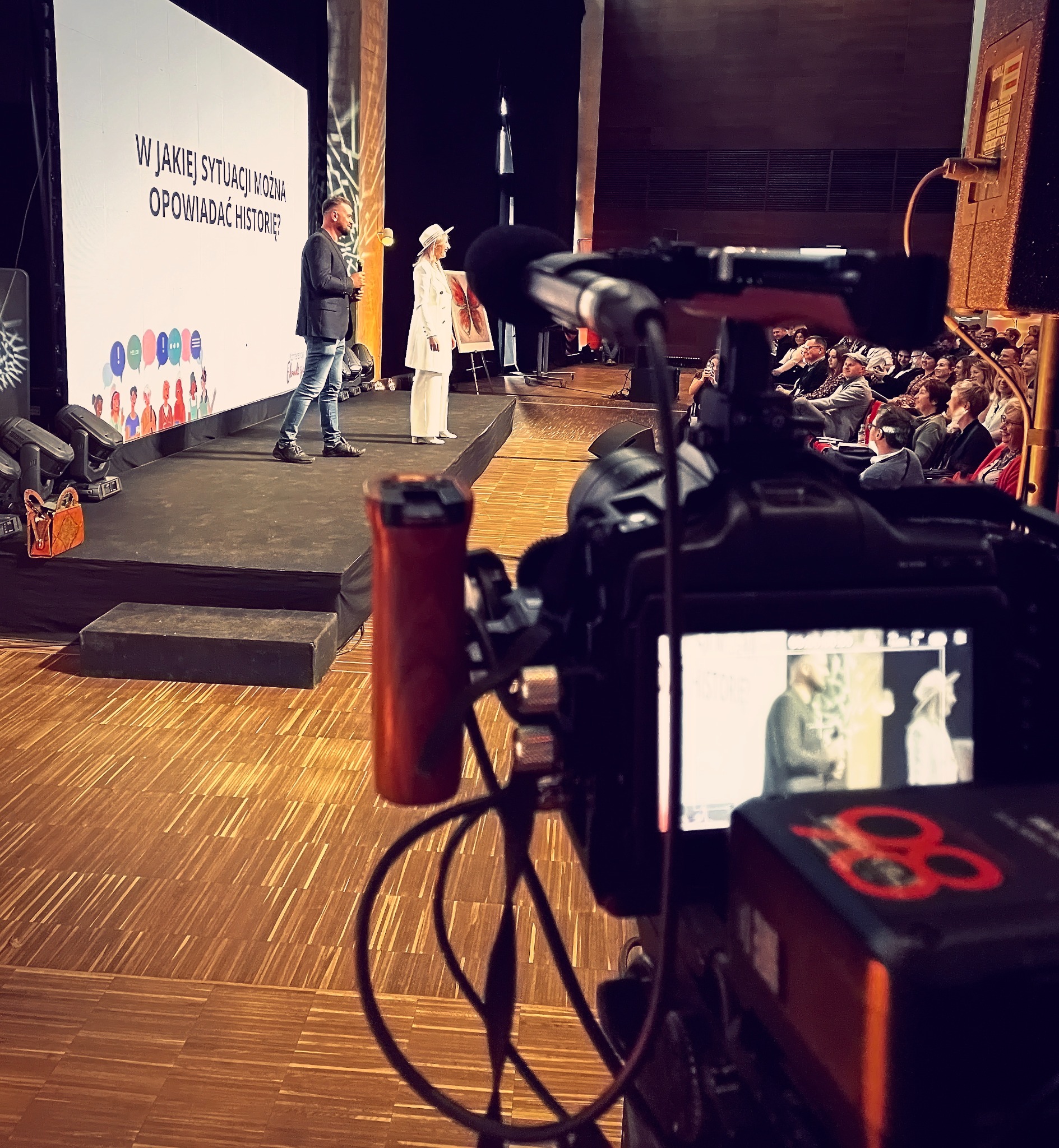 Kamera nagrywająca prelekcję dwóch osób na scenie z publicznością w tle, ekran z napisem 'W jakiej sytuacji można opowiadać historie?'