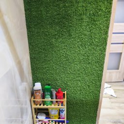 SebBud - Ściana w łazience wyłożona sztuczną trawą, obok półka z kosmetykami. Nietypowe wykończenie wnętrza z akcentem zieleni i naturalnego drewna.