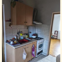 SebBud - Wnętrze kuchni przed remontem: widok na zabudowę kuchenną z czerwonym zlewem, kuchenkę gazową i płytki na ścianach. Widoczny bałagan i zużycie.