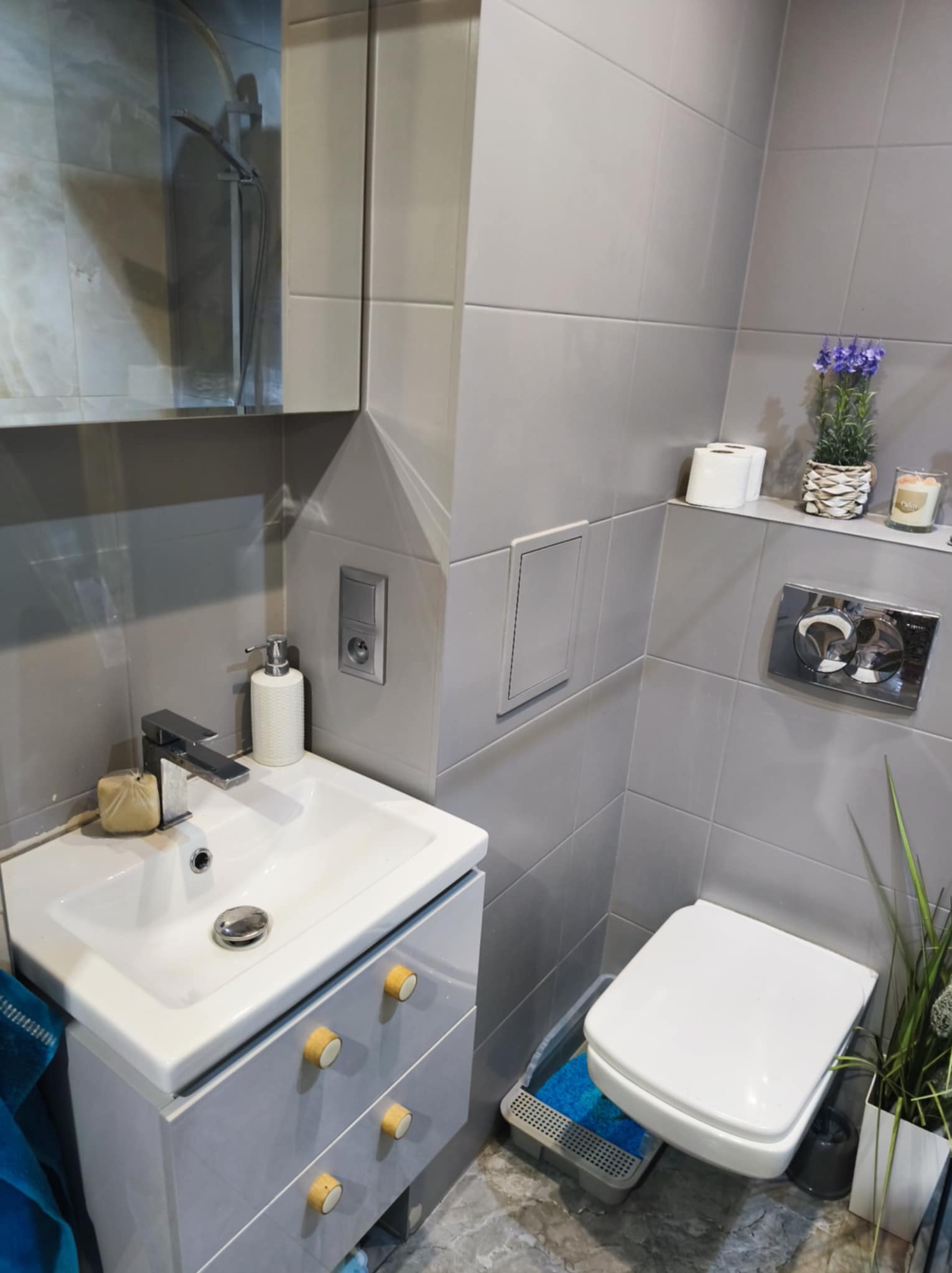 Nowoczesna łazienka w szarościach: umywalka z szafką z drewnianymi uchwytami, toaleta, płytki ceramiczne i dekoracyjne akcenty roślinne.