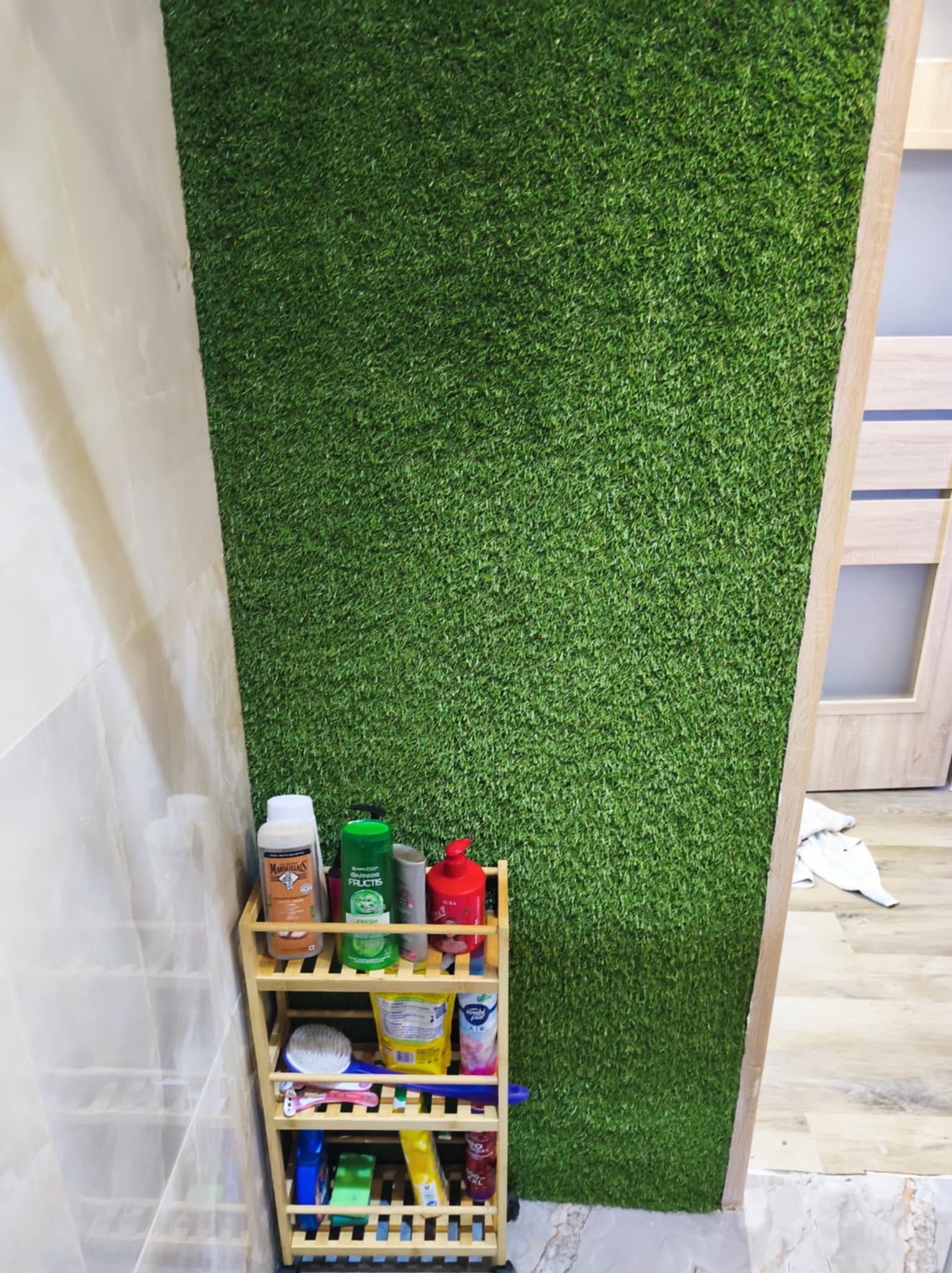 Ściana w łazience wyłożona sztuczną trawą, obok półka z kosmetykami. Nietypowe wykończenie wnętrza z akcentem zieleni i naturalnego drewna.