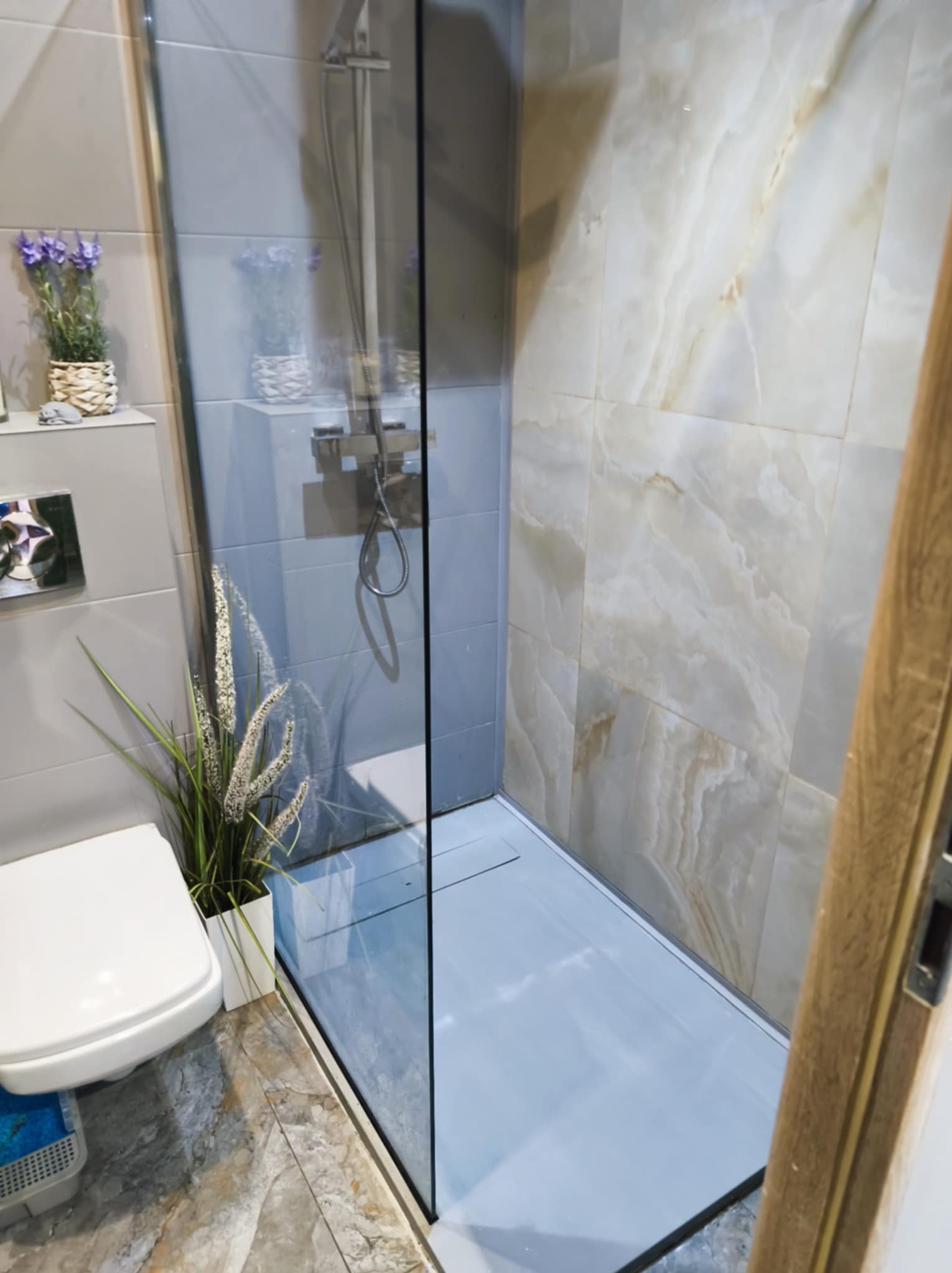 Wykończona łazienka z kabiną prysznicową typu walk-in, marmurowe płytki na ścianach, toaleta i dekoracyjne rośliny. Minimalistyczny design z akcentami natury.