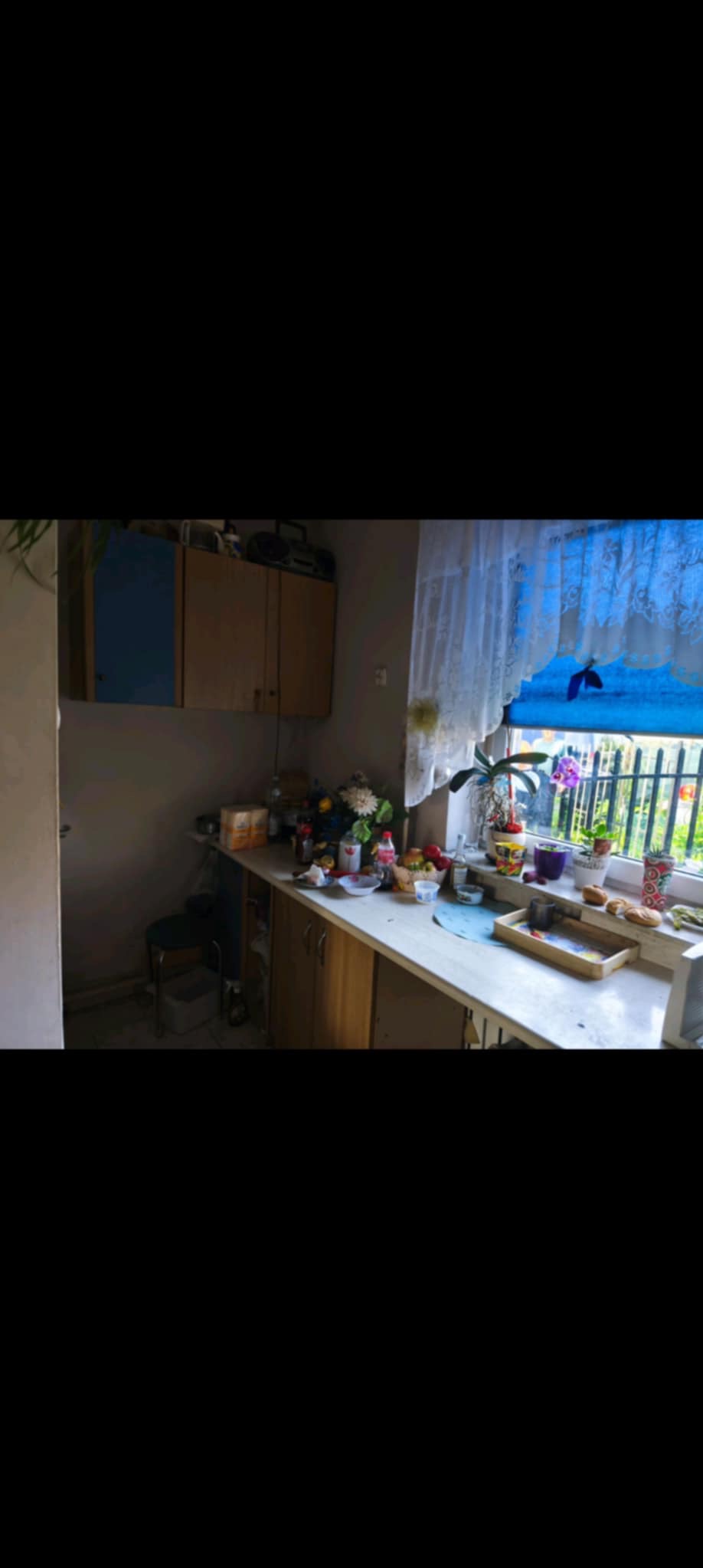 Kuchnia przed remontem: widok na blat z oknem, szafki wiszące i podłogowe. Na blacie kwiaty i akcesoria kuchenne. Ściany szare, wymagające odświeżenia.