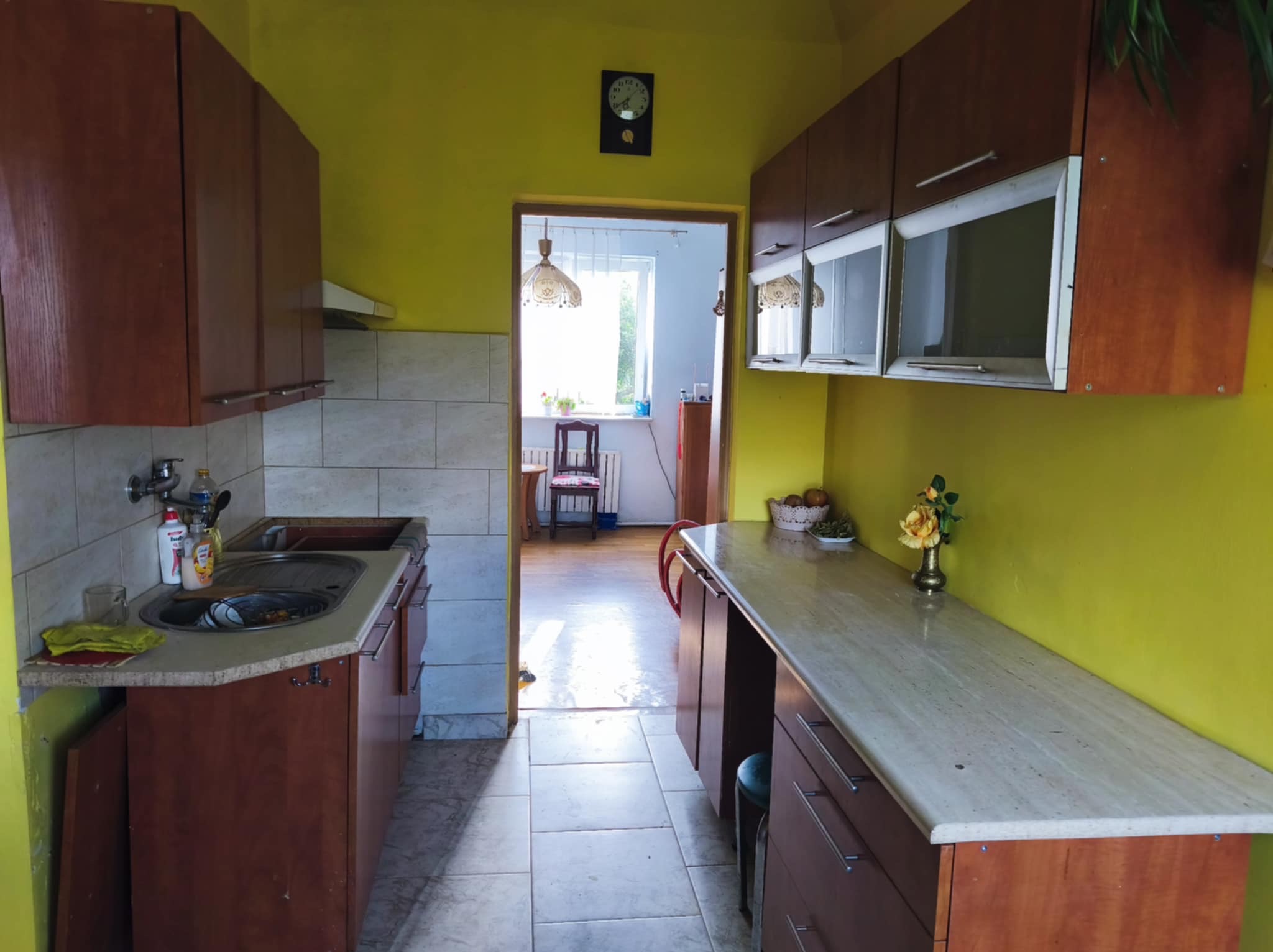 Kuchnia z żółtymi ścianami, brązowymi szafkami i marmurowym blatem. Widoczny zlew, okap i zegar na ścianie. W tle widoczne inne pomieszczenie z krzesłem i lampą.