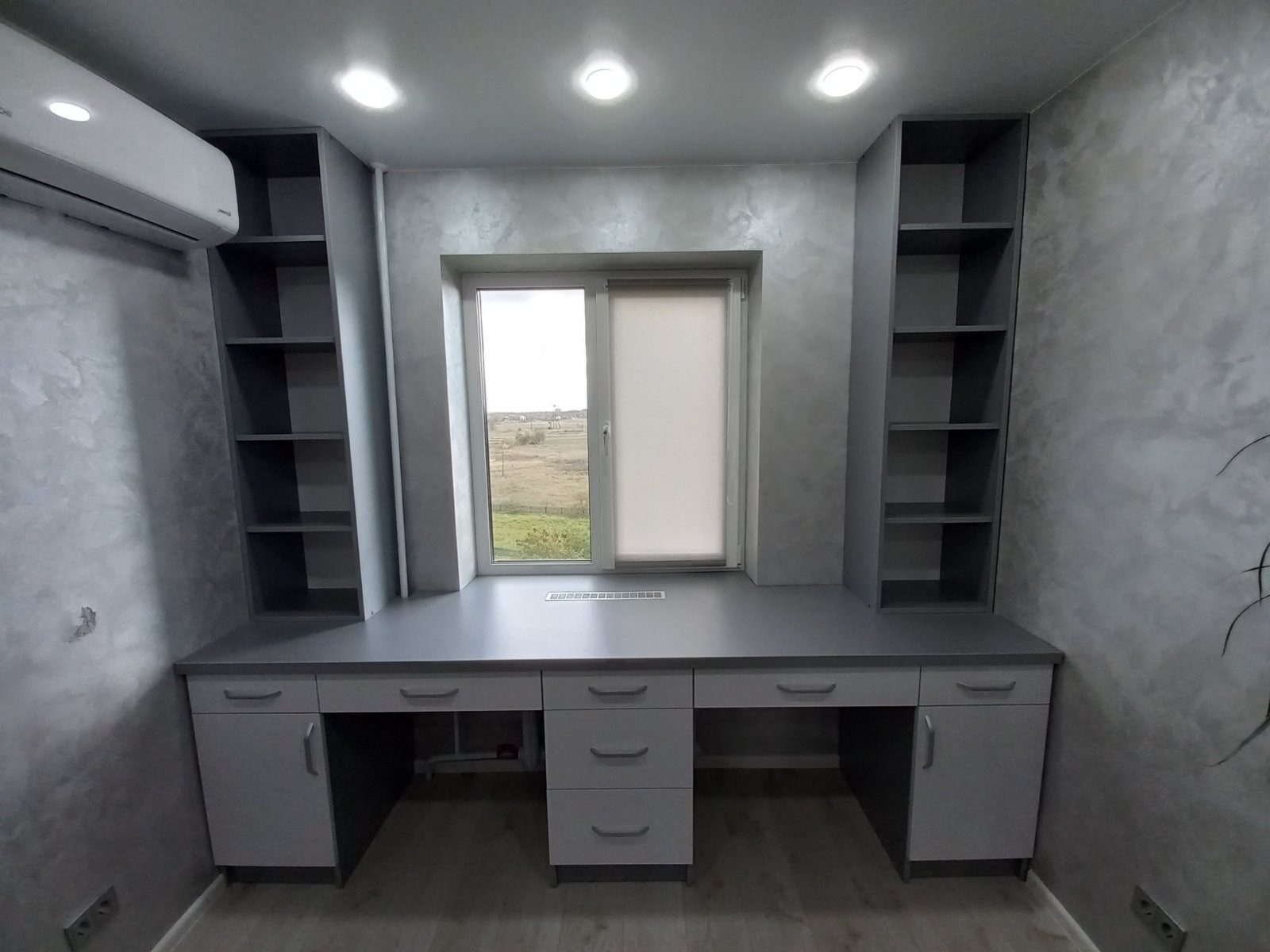 Szara zabudowa meblowa z biurkiem i regałami w pokoju z gładzią dekoracyjną, widok na okno z roletą. Oświetlenie sufitowe punktowe.