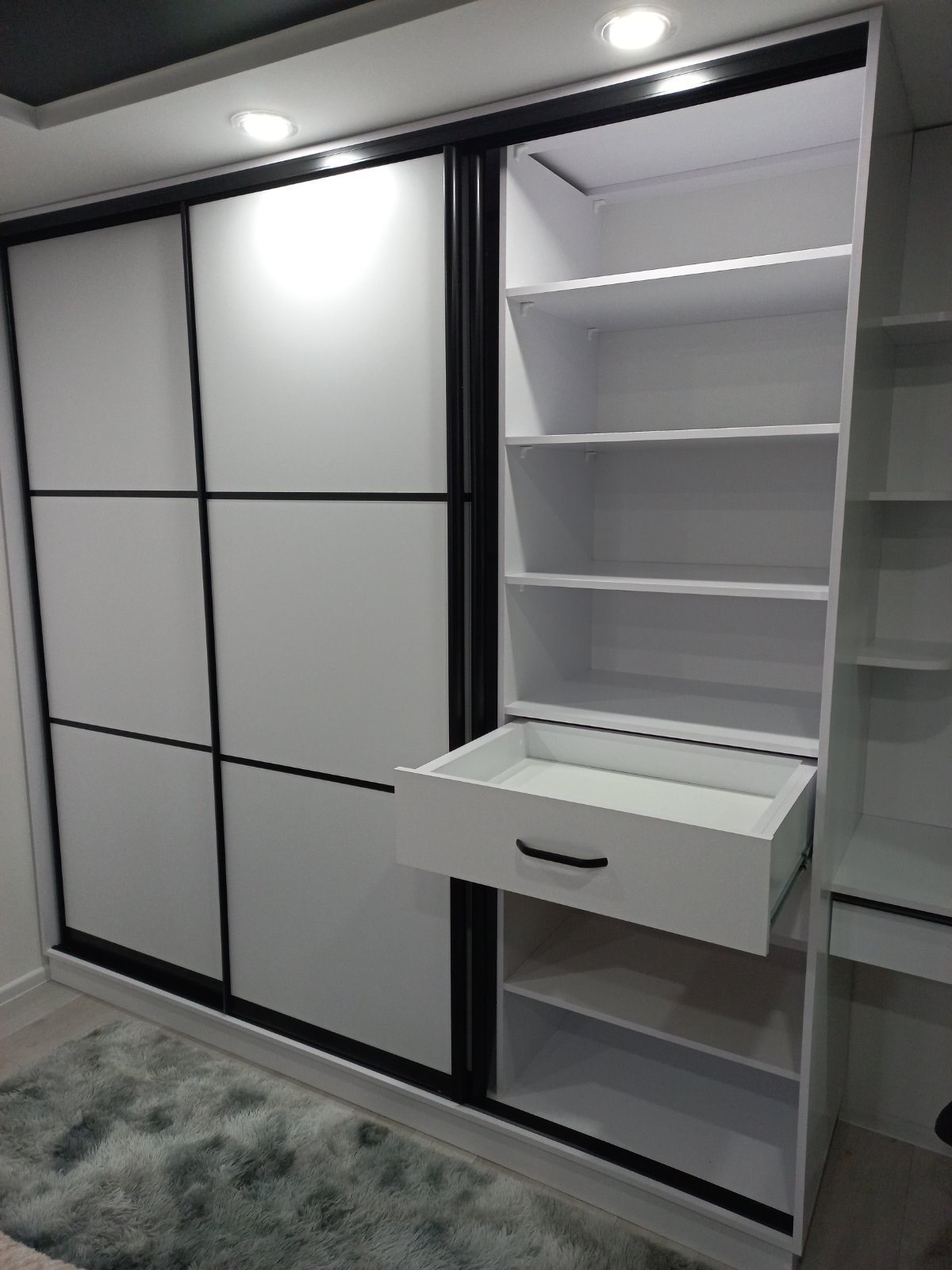 Biała szafa wnękowa z przesuwnymi drzwiami i czarnymi ramami, z wysuniętą szufladą, oświetleniem punktowym i miękkim dywanem w tle.