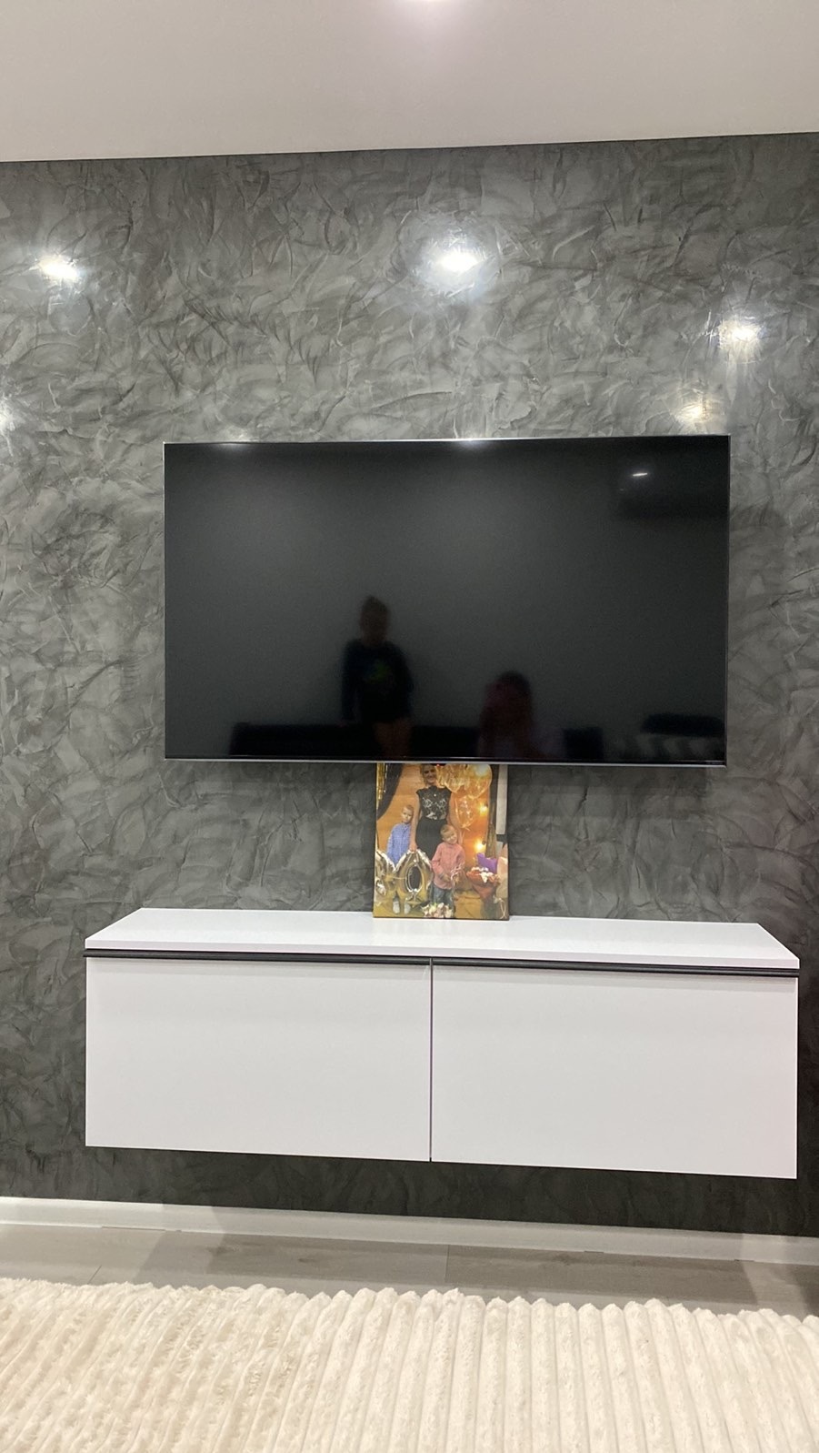 Ściana z efektem dekoracyjnym w odcieniach szarości, podwieszana szafka RTV z obrazem i wiszący telewizor. Nowoczesny design wnętrza.