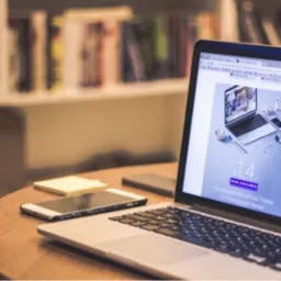 Laptop z otwartym projektem interfejsu strony internetowej na ekranie, obok telefon, notatki i długopis na drewnianym biurku, w tle regał z książkami.