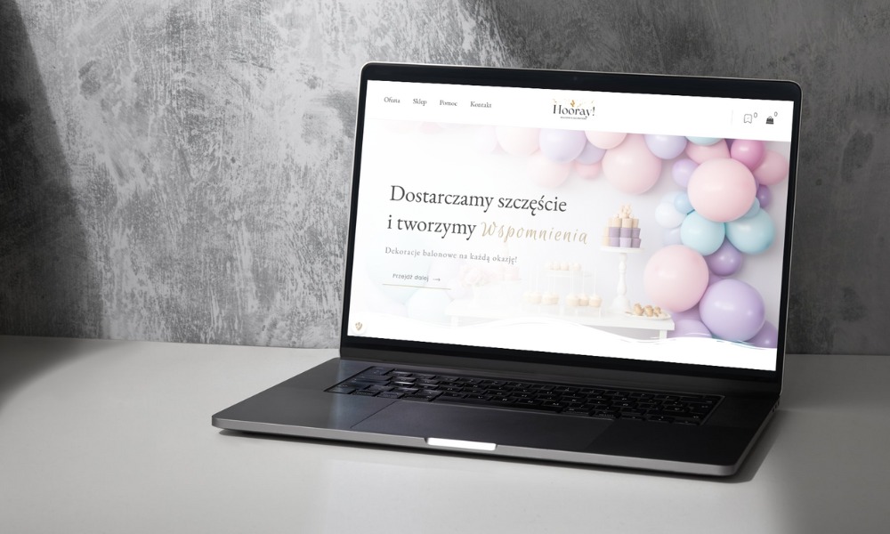 Laptop wyświetlający stronę internetową z dekoracjami balonowymi na tle szarej, teksturowanej ściany. Strona zawiera logo 'Hooray!' i hasło 'Dostarczamy szczęście i tworzymy Wspomnienia'.