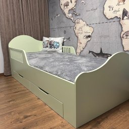 FORMHAUS Michał Ozga - Łóżko dziecięce w kolorze pistacjowym z szufladami, zdobione gwiazdkami i księżycem, z poduszką z motywem pojazdów budowlanych, na tle ściany z mapą świata.