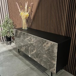 FORMHAUS Michał Ozga - Komoda z frontami imitującymi marmur, z czarnym blatem, na tle ściany z drewnianych paneli i zasłony, z dekoracjami roślinnymi na blacie i obok.