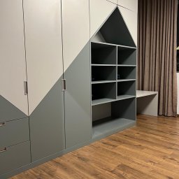 FORMHAUS Michał Ozga - Dwukolorowa szafa wnękowa z elementami w kształcie domku i otwartymi półkami, uzupełniona blatem roboczym, na tle brązowych zasłon i drewnianej podłogi.