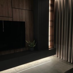 FORMHAUS Michał Ozga - Nowoczesna zabudowa ściany telewizyjnej z elementami drewnopodobnymi, pionowymi lamelami i podświetlanymi półkami, uzupełniona szarymi zasłonami i podświetleniem podłogowym.