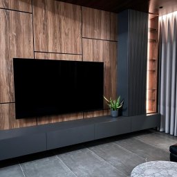 FORMHAUS Michał Ozga - Nowoczesna ściana telewizyjna z drewnianymi panelami, czarnym telewizorem, szarą szafką RTV i podświetlaną półką, uzupełniona zieloną rośliną w doniczce.