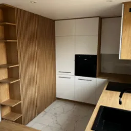 Nowoczesna kuchnia z białymi frontami szafek, czarnymi uchwytami i piekarnikiem w zabudowie, drewnianym blatem oraz ścianą z drewnianych listew i półkami.