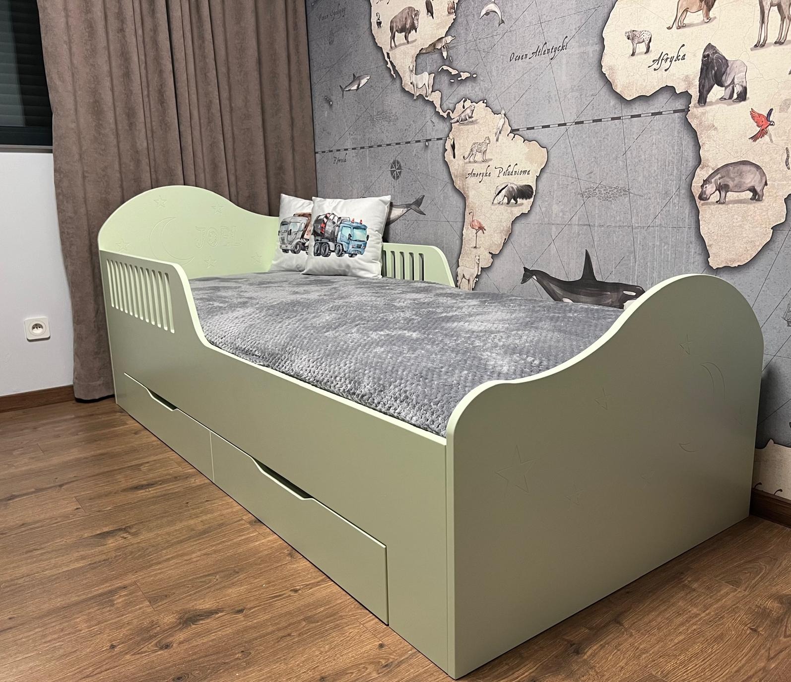 Łóżko dziecięce w kolorze pistacjowym z szufladami, zdobione gwiazdkami i księżycem, z poduszką z motywem pojazdów budowlanych, na tle ściany z mapą świata.