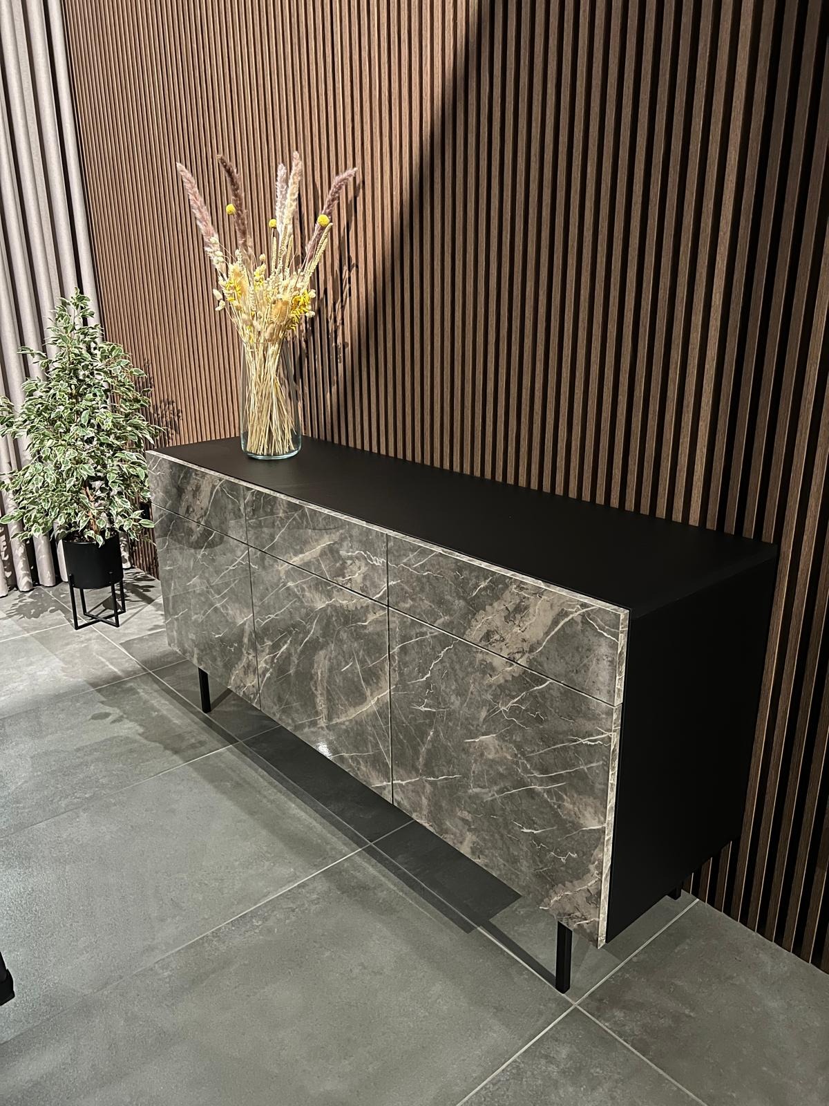 Komoda z frontami imitującymi marmur, z czarnym blatem, na tle ściany z drewnianych paneli i zasłony, z dekoracjami roślinnymi na blacie i obok.