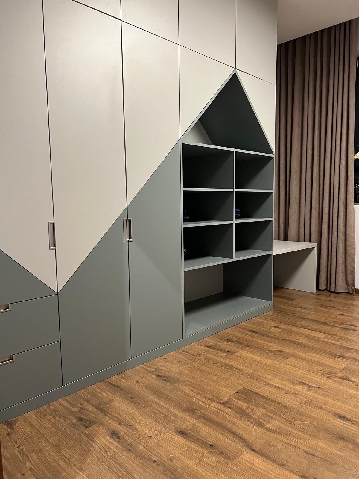 Dwukolorowa szafa wnękowa z elementami w kształcie domku i otwartymi półkami, uzupełniona blatem roboczym, na tle brązowych zasłon i drewnianej podłogi.