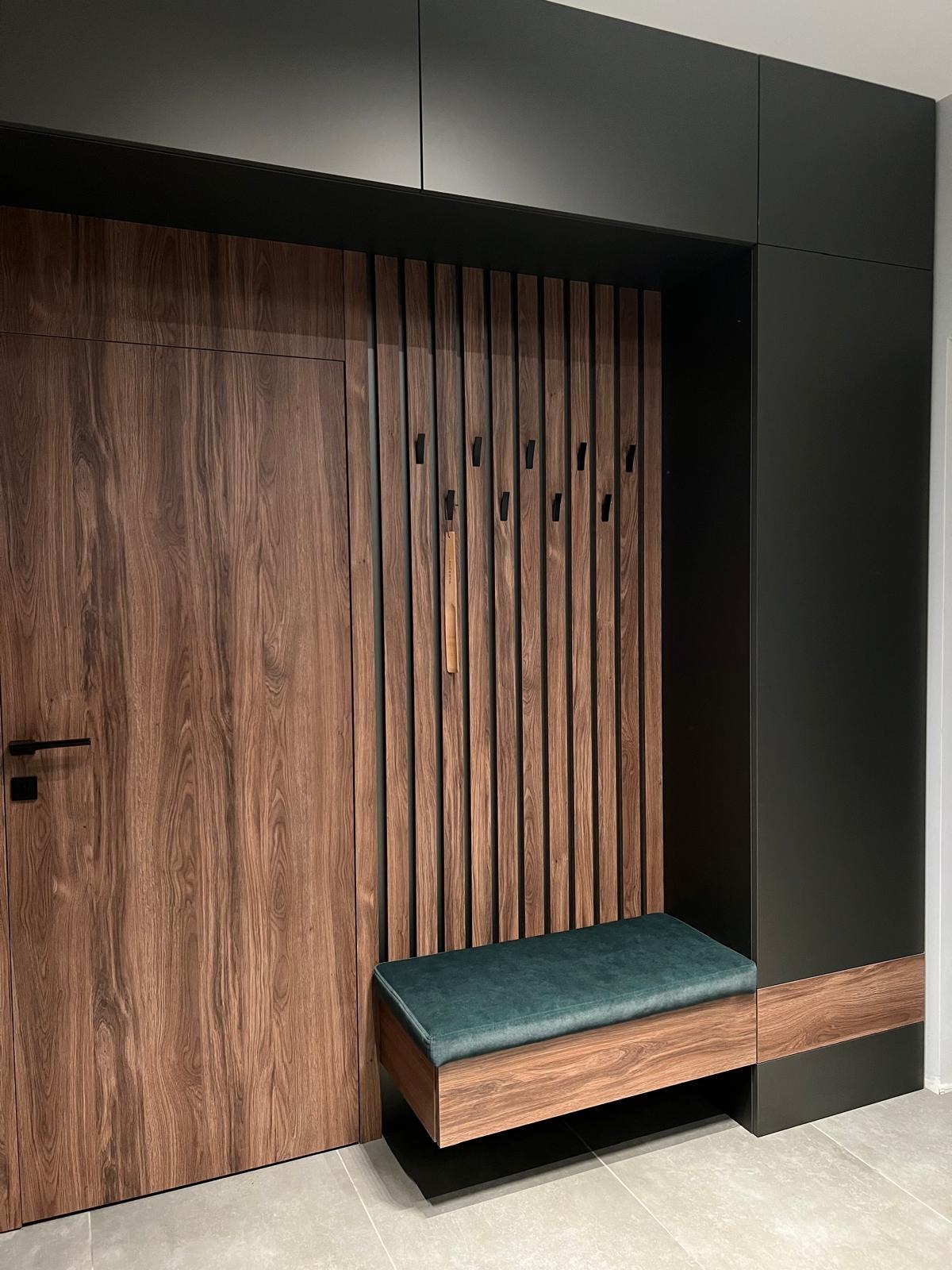 Nowoczesna zabudowa przedpokoju z drewnianymi lamelami, wieszakami i zielonym siedziskiem, całość w stonowanej kolorystyce.